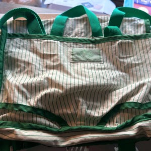 May include: A canvas tote bag with a cream and green striped pattern. The bag features green trim, handles, and shoulder straps. A small green label is sewn onto the front. The bag is designed for carrying items.