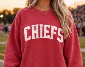 May include: A red crewneck sweatshirt featuring the word CHIEFS printed in a distressed white font across the chest. The garment has long sleeves with ribbed cuffs and a ribbed hem, providing a relaxed fit with a classic athletic appearance.