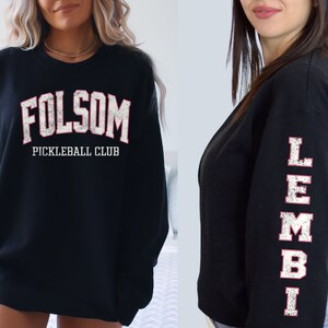 May include: Two black sweatshirts with white and pink lettering. One sweatshirt says "Folsom Pickleball Club" and the other has "Lembi" written vertically on the sleeve.