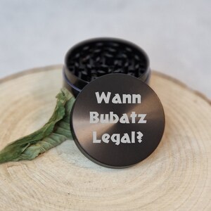 Puede incluir: Un molinillo de hierbas negro con la tapa abierta, revelando los dientes de moler. La tapa tiene el texto "Wann Bubatz Legal?" en blanco. El molinillo está sobre una superficie de madera con una hoja verde.