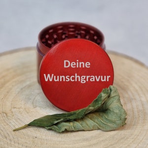 Op de afbeelding: Rode metalen grinder met een deksel waarop "Deine Wunschgravur" in witte letters staat. De grinder staat op een houten oppervlak met een droog blad.