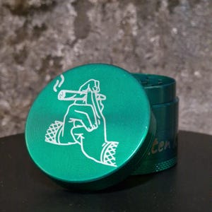 Puede incluir: Un molinillo de metal verde con un diseño blanco de una mano sosteniendo un cigarrillo. El molinillo tiene el texto "Green Room" en el lateral.