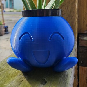 Op de afbeelding: Een blauwe 3D-geprinte plantenpot in de vorm van een lachend cartoonfiguur met een kleine aloëplant die erin groeit.