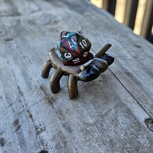 May include: A 3D printed, brown and black spider with six legs holding a multi-colored, 20-sided die with white numbers.