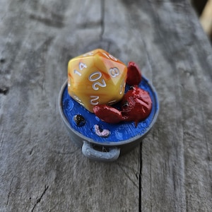 May include: A yellow and orange 20-sided die with white numbers sits in a blue pot with a red crab. The pot has a gray handle.