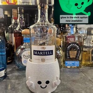 May include: White bottle holder with a smiling face and two raised middle fingers. The holder is holding a bottle of Martell Cognac. The bottle holder is on a bar counter with other bottles of alcohol.