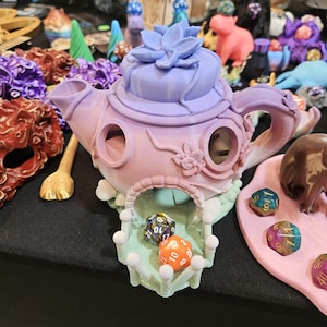 May include: A pastel purple and pink teapot shaped dice tower with a flower on top. The tower has a small opening for dice to roll out. There are four dice in the tower, two blue, one orange, and one multi-colored.