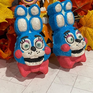 May include: Two blue bunny-shaped candies with white accents on their ears and heads, pink cheeks and bows, and black mouths. The candies are in front of a Halloween-themed container with candy and fall leaves.