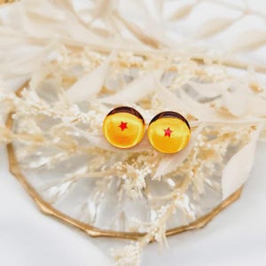 May include: A pair of gold-colored stud earrings. Each earring features a transparent, spherical design with a red star in the center and a dark brown top. The earrings are set against a backdrop of dried flowers and a gold-edged tray.