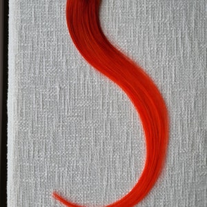 Puede incluir: Una extensión de cabello naranja vibrante, curvada en forma fluida, descansa sobre una superficie blanca texturizada. La extensión es de un color brillante y sólido, y parece estar hecha de fibras sintéticas. El borde superior muestra una trama para la fijación.