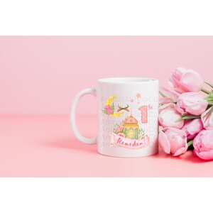 May include: White ceramic mug with a pink and yellow Ramadan design. The design features a crescent moon, a mosque, a lantern, and the words "1st Ramadan".