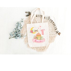 May include: A white canvas tote bag with a pink and yellow design featuring a crescent moon, a star, a mosque, a lantern, and the text "1st Ramadan".