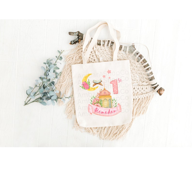 May include: A white canvas tote bag with a pink and yellow design featuring a crescent moon, a star, a mosque, a lantern, and the text "1st Ramadan".