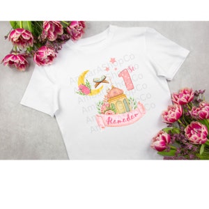 May include: A white t-shirt with a pink and yellow design featuring a crescent moon, a lantern, and the number "1" with the word "Ramadan" on a pink ribbon.