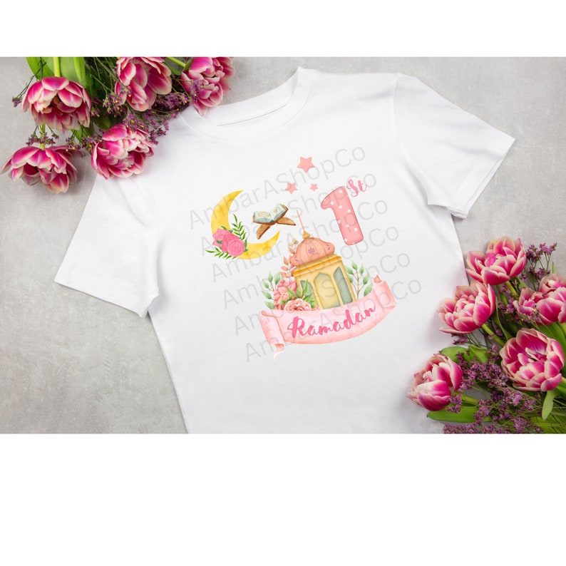 May include: A white t-shirt with a pink and yellow design featuring a crescent moon, a lantern, and the number "1" with the word "Ramadan" on a pink ribbon.