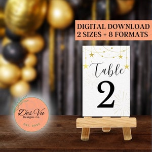 Puede incluir: Tarjeta de número de mesa dorada con fondo blanco y estrellas doradas. La tarjeta dice "Mesa 2" y se muestra en un pequeño caballete de madera. La tarjeta es parte de un conjunto de números de mesa para una fiesta o evento.