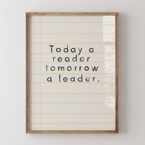 Op de afbeelding: Een ingelijste print met de tekst "Today a reader tomorrow a leader" in zwarte letters. De achtergrond lijkt op gelijnd papier met gele lijnen. Het frame is gemaakt van lichtbruin hout.