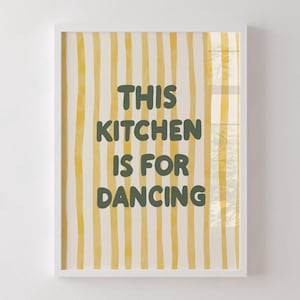 Op de afbeelding: Wit ingelijste kunstdruk met een crèmekleurige achtergrond en verticale gele strepen. De tekst "THIS KITCHEN IS FOR DANCING" staat in donkergroene, speelse letters. Vrolijke keukenwanddecoratie.