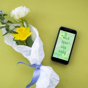 May include: A bouquet of white and yellow flowers wrapped in white tissue paper with a blue ribbon. A smartphone with a green screen displaying the text "WILD FREE AND ADHD" is lying on a green surface.