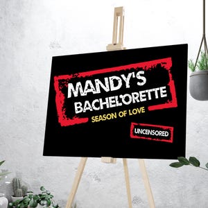 Könnte beinhalten: Ein schwarzes Schild auf einer Staffelei mit dem Text "MANDY'S BACHELORETTE SEASON OF LOVE" in Weiß und Rot. Das Wort "UNCENSORED" wird ebenfalls in einem roten Feld angezeigt. Das Schild wird vor einer weißen Wand angezeigt.