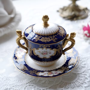 Könnte beinhalten: Ein kobaltblaues und goldfarbenes Porzellan-Tee-Set. Die Tasse hat einen Deckel und verzierte goldene Griffe. Das Set zeigt florale Designs und goldene Filigranakzente. Die Untertasse ist rund und passt zum Design der Tasse.