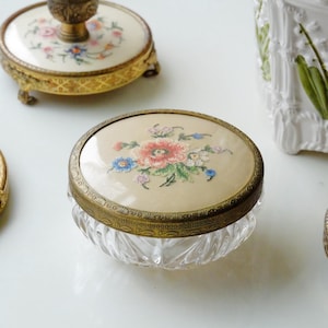 Puede incluir: Un tarro de cristal vintage con una tapa bordada con flores. La tapa presenta una tela color crema con una flor roja central rodeada de flores azules y blancas. El tarro tiene un borde de latón decorativo y una base de cristal transparente facetada. Otros artículos antiguos están en el fondo.