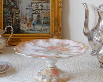 Może przedstawiać: Biały porcelanowy stojak na ciasto z falowanym brzegiem i kwiatowym wzorem. Stojak zdobią różowe i żółte róże z zielonymi liśćmi i złotymi akcentami. Stojak stoi na białej koronkowej serwetce.