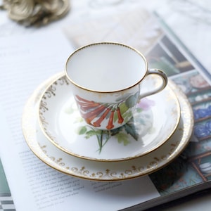 Puede incluir: Un juego de taza, platillo y plato de porcelana blanca. La taza y los platos tienen ribetes dorados y un diseño floral con detalles en rojo, verde y morado. El juego está sobre una revista.