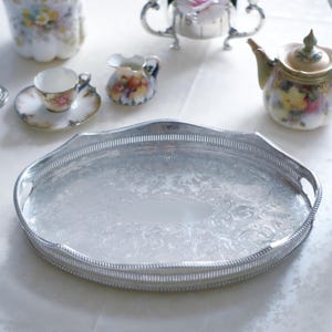May include: A silver oval serving tray with a decorative floral pattern and a beaded edge. A tea set is arranged around it, including a teapot, creamer, cup, and saucer, all with floral designs.