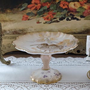 Könnte beinhalten: Ein weißer Porzellan-Kuchenständer mit einem gewellten Rand und einem Blumendesign. Der Ständer zeigt ein Paar Vögel, die auf einem Zweig mit Blumen sitzen. Der Ständer hat einen goldenen Rand und einen rosa Sockel.