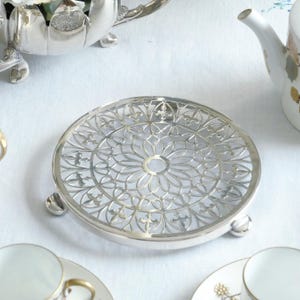 Puede incluir: Un salvamanteles redondo plateado con un intrincado diseño calado. El salvamanteles tiene un patrón floral en el centro y está soportado por tres pequeños pies. Está diseñado para proteger las superficies de teteras o platos calientes.
