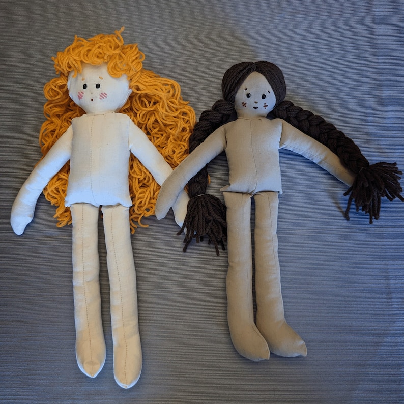 May include: Two handmade fabric dolls with yarn hair. The doll on the left has blonde yarn hair and a white dress. The doll on the right has brown yarn hair and a gray dress.