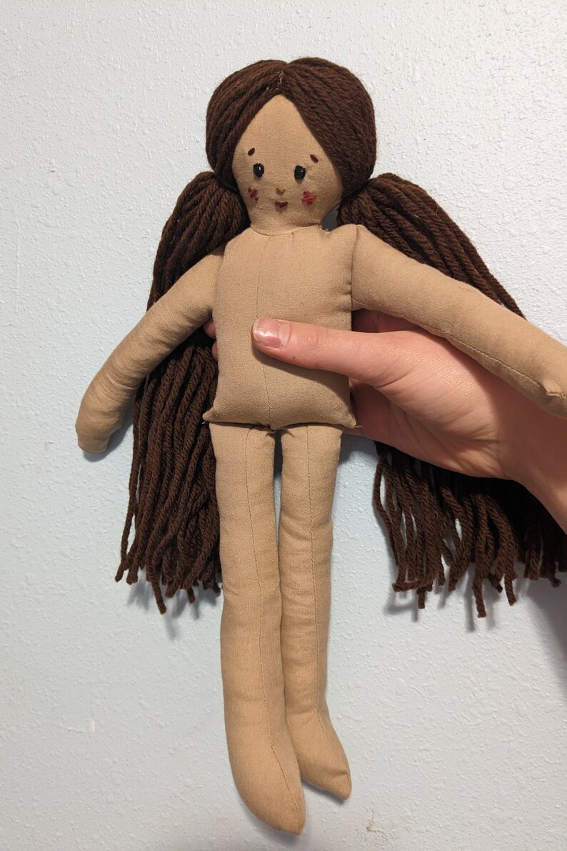 May include: A handmade rag doll with brown yarn hair, a beige fabric body, and a stitched face with brown eyes and red cheeks.