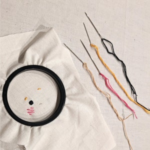 May include: A white embroidery hoop with a black embroidery frame. The hoop is on a white fabric surface. There are four embroidery needles with different colored threads attached to them.