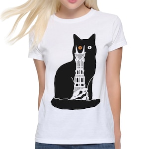 Puede incluir: Camiseta blanca con una silueta negra de un gato con un ojo naranja y un ojo blanco. El gato sostiene una torre negra y blanca en sus patas.