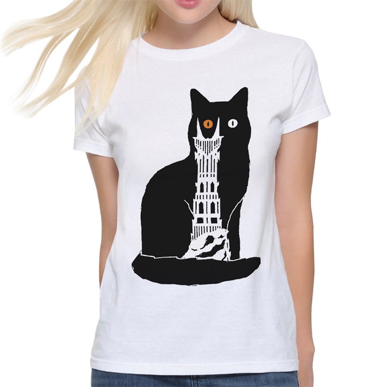 Puede incluir: Camiseta blanca con una silueta negra de un gato con un ojo naranja y un ojo blanco. El gato sostiene una torre negra y blanca en sus patas.