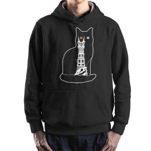 May include: Black hooded sweatshirt featuring a white outline of a cat with a tower design inside. The cat has an orange eye. The sweatshirt has a front pocket and a drawstring hood.