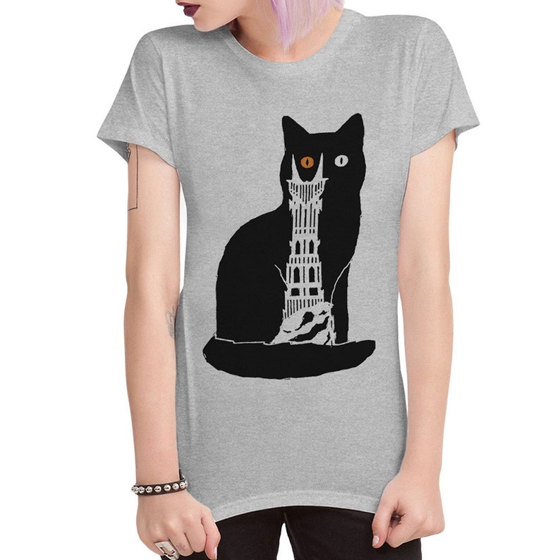 Puede incluir: Una camiseta gris con una silueta de gato negro. El gato tiene un ojo naranja y un ojo blanco. El cuerpo del gato est&aacute; lleno de una silueta negra y blanca de una torre.
