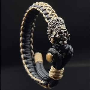 Puede incluir: Pulsera artesanal con un colgante de calavera detallado con un tocado ornamentado. La pulsera está hecha de cordón trenzado negro y beige, con un acento dorado. El colgante de calavera es de color metálico oscuro.