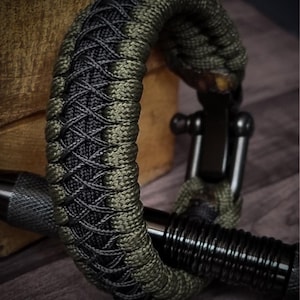 May include: A close-up of a braided bracelet. The bracelet is made of olive green and black cord, with a black metal clasp. The bracelet is resting on a wooden surface. The bracelet is a stylish accessory.