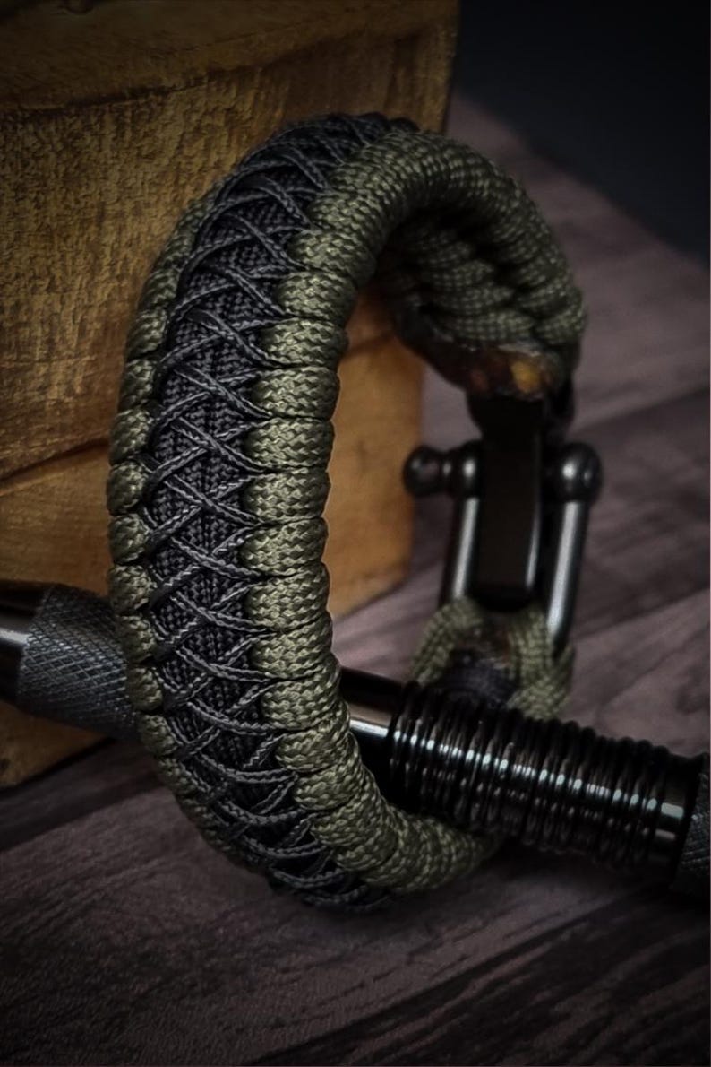 May include: A close-up of a braided bracelet. The bracelet is made of olive green and black cord, with a black metal clasp. The bracelet is resting on a wooden surface. The bracelet is a stylish accessory.
