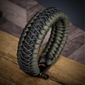 May include: A braided bracelet in shades of green and black, resting on a wooden surface. The bracelet features a complex woven pattern and a black metal clasp. The design is intricate and the color combination is striking.