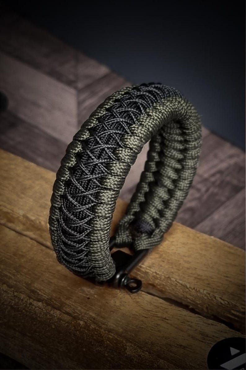 May include: A braided bracelet in shades of green and black, resting on a wooden surface. The bracelet features a complex woven pattern and a black metal clasp. The design is intricate and the color combination is striking.
