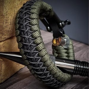 May include: A close-up of a bracelet with a woven design. The bracelet features a combination of olive green and gray paracord, with a black metal clasp. The design is intricate and detailed, showcasing the craftsmanship of the piece.