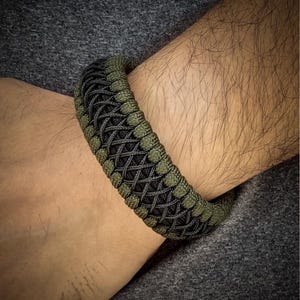 May include: A woven bracelet in shades of olive green and black, worn on a wrist. The bracelet features a complex, textured pattern. The background is a dark gray fabric.