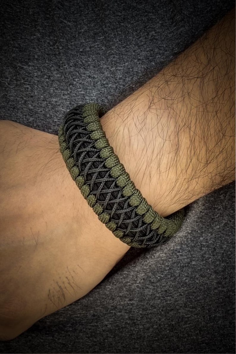 May include: A woven bracelet in shades of olive green and black, worn on a wrist. The bracelet features a complex, textured pattern. The background is a dark gray fabric.