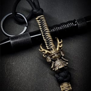Könnte beinhalten: Ein detaillierter Schlüsselanhänger mit einem Miniatur-Samuraischwert-Anhänger. Das Schwert hat einen bronzefarbenen Griff und einen schwarz-goldenen geflochtenen Griff. Der Schlüsselanhänger enthält eine schwarze Kordelschlaufe und einen schwarzen Metallring.