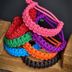 May include: A stack of colorful paracord bracelets in pink, red, green, blue, orange, and black. The bracelets are made with a woven knot pattern and are stacked on a wooden surface.