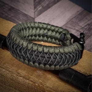 May include: A close-up of a paracord bracelet in shades of green and black. The bracelet is intricately woven with a textured pattern. The bracelet is resting on a wooden surface, with a black metal clasp and a black flashlight visible.