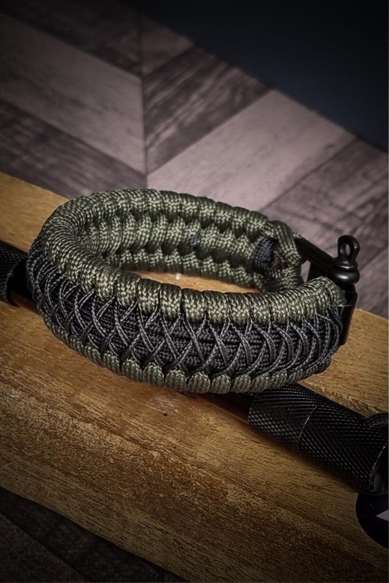 May include: A close-up of a paracord bracelet in shades of green and black. The bracelet is intricately woven with a textured pattern. The bracelet is resting on a wooden surface, with a black metal clasp and a black flashlight visible.